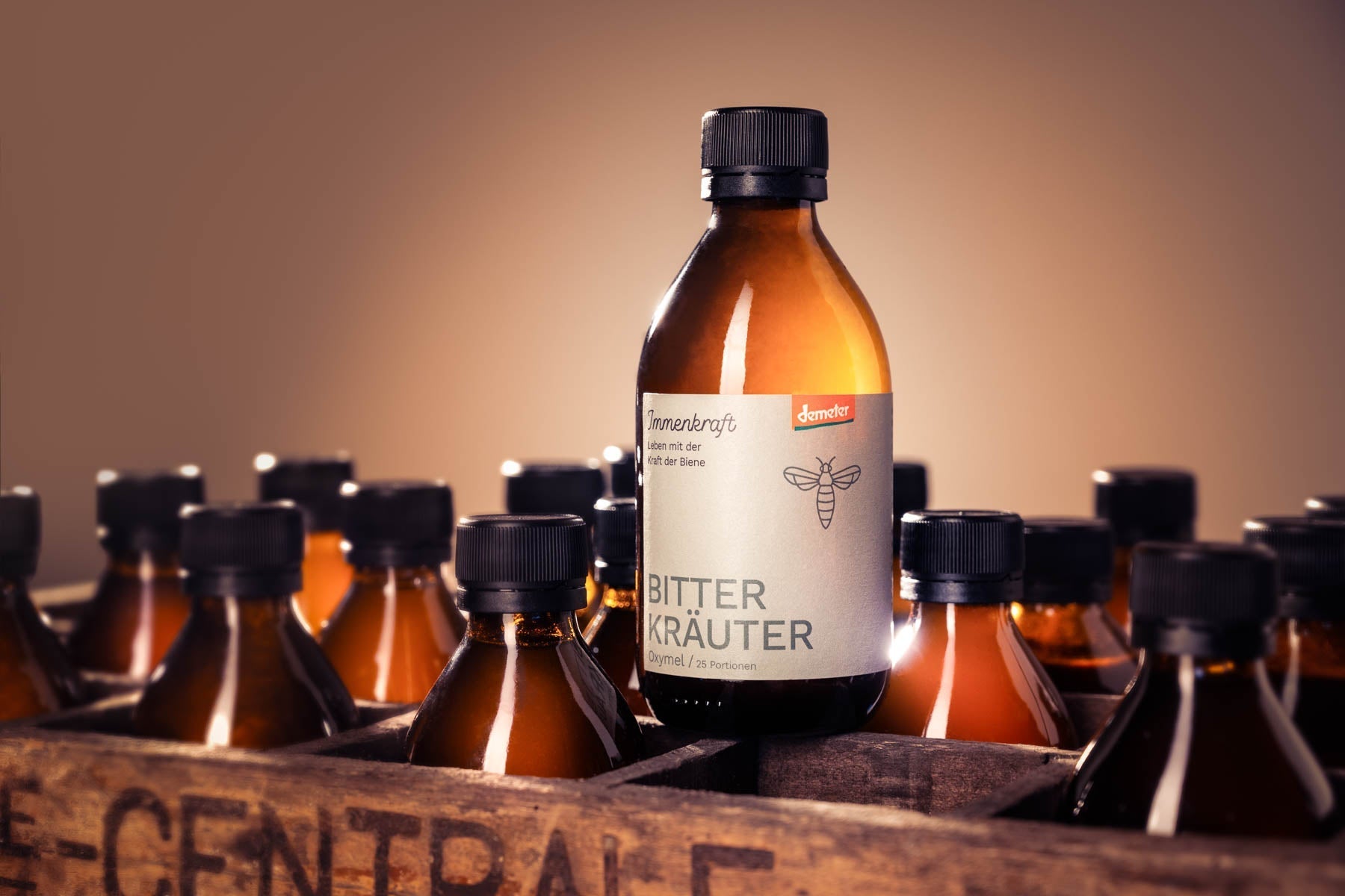 Eine antike Limonadenkiste aus Holz in welchem Immenkraft Demeter-Oxymel Flaschen in verschiedenen Sorten stehen. Oben eine Flasche Immenkraft Bitterkräuter Demeter-Oxymel.