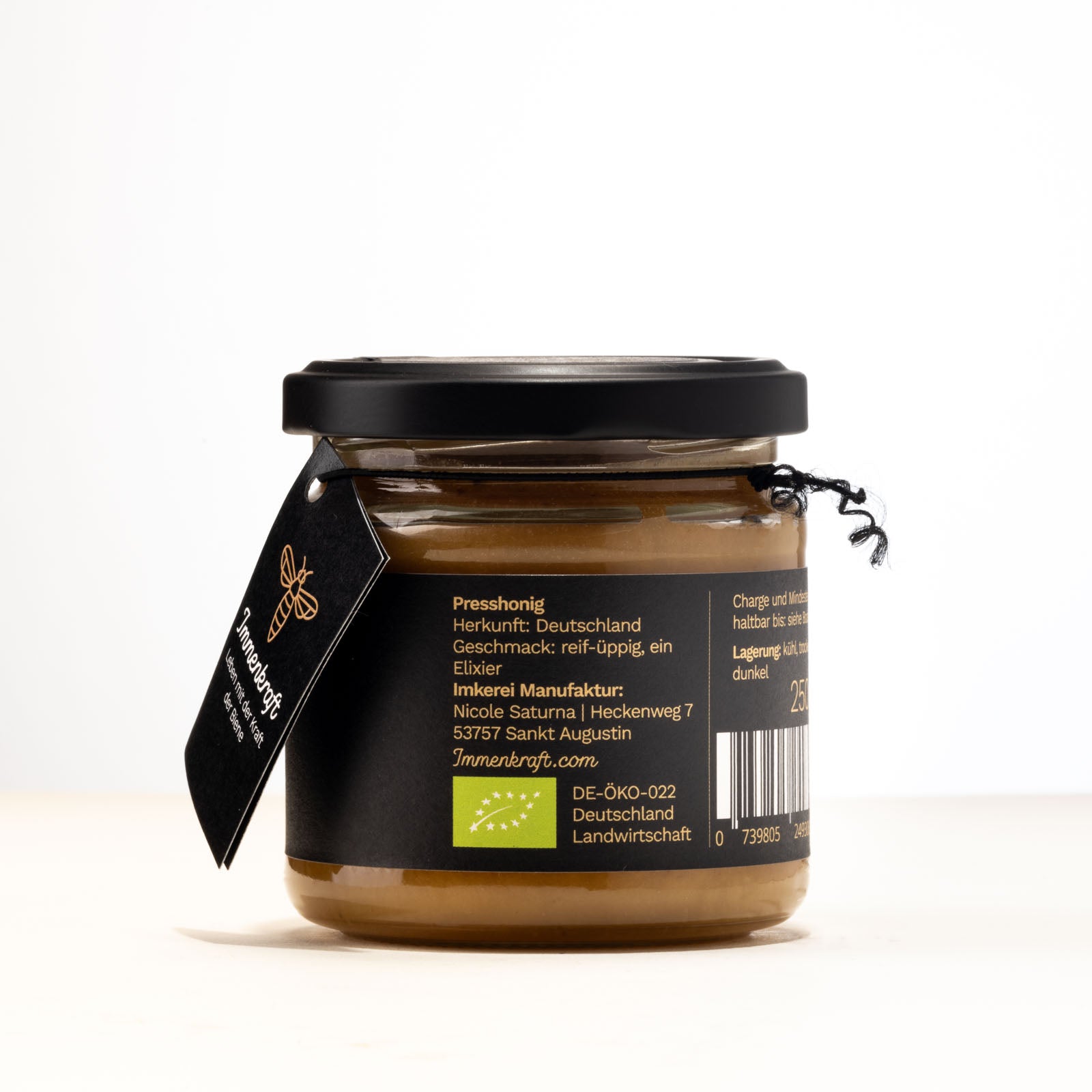 Ein 250g klares Twist-Off-Glas mit matt-schwarzem Deckel, schwarzem Anhängeetikett und Etikett mit der Aufschrift Immenkraft - Leben mit der Kraft der Biene. Produktname: Quintessenz Presshonig. Seitenansicht: Herkunft, Herstelleradresse, Geschmack und EU-Bio-Logo.