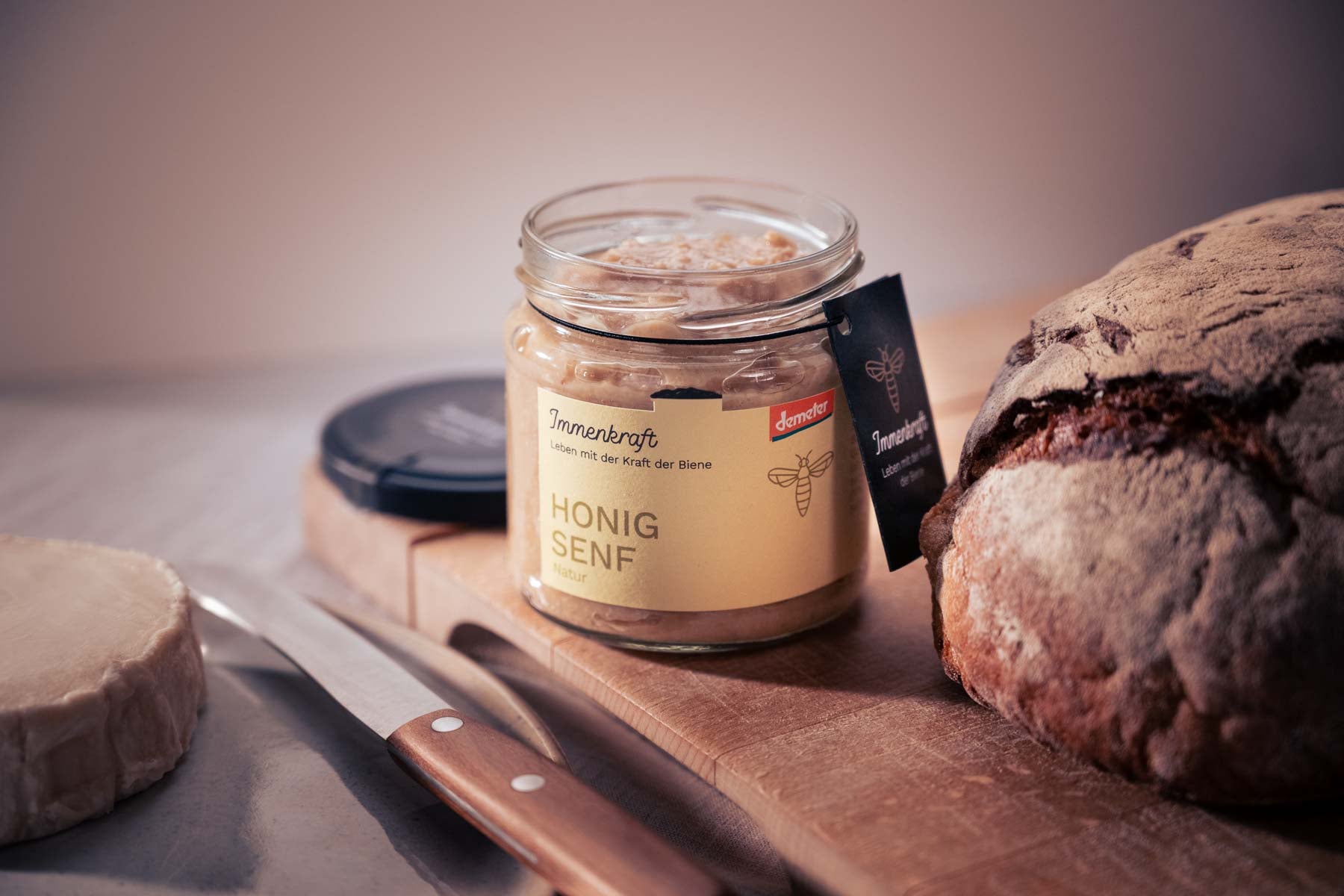 Anwendungsbeispiel für Demeter-Honigsenf, Natur: Auf einem rustikalen Holzbrett steht ein geöffnetes Glas Immenkraft Honigsenf, daneben ein schönes, dunkles Brot und davor ein Manufaktur-Brotmesser und ein Stück Ziegenkäse