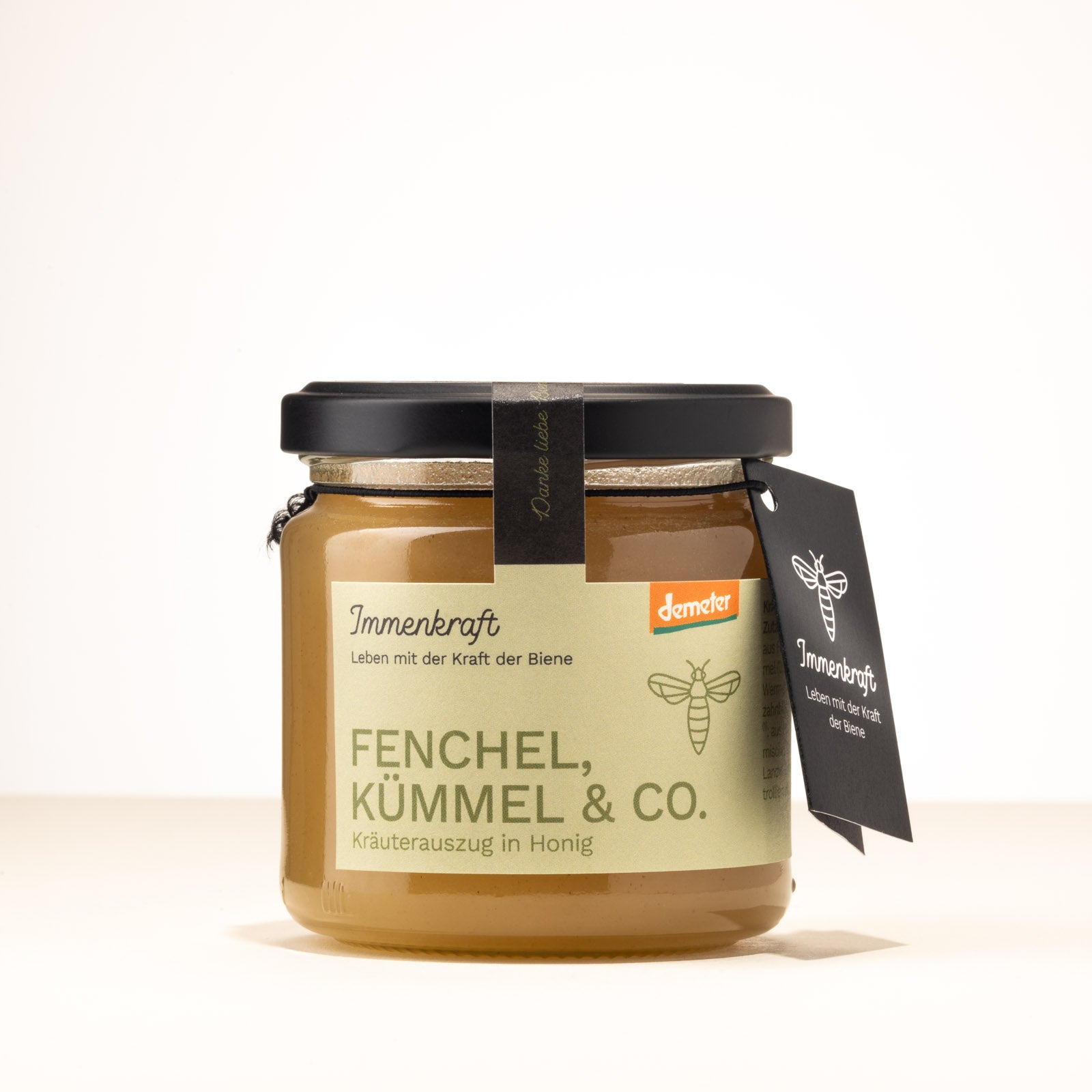 Ein 250g klares Twist-Off-Glas mit matt-schwarzem Deckel, schwarzem Anhängeetikett und Etikett mit der Aufschrift Immenkraft - Leben mit der Kraft der Biene. Produktname: Fenchel, Kümmel & Co., Kräuterauszug in Honig. Demeter-Siegel und Bienen-Logo.