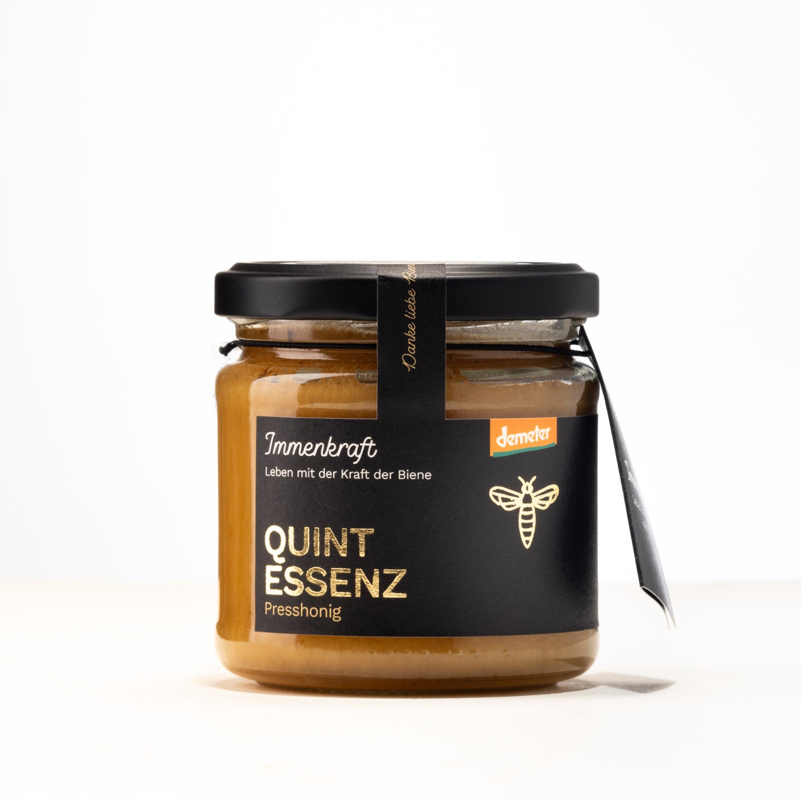 Ein 250g klares Twist-Off-Glas mit matt-schwarzem Deckel, schwarzem Anhängeetikett und Etikett mit der Aufschrift Immenkraft - Leben mit der Kraft der Biene. Produktname: Quintessenz-Presshonig. Demeter-Siegel und Bienen-Logo.