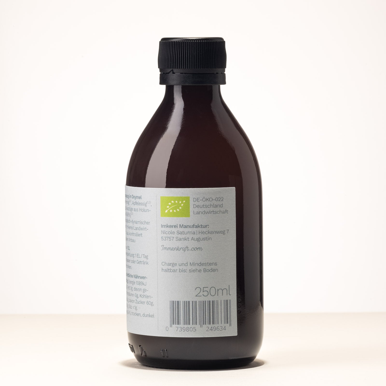 Eine Braunglas-Flasche mit der Aufschrift Immenkraft - Leben mit der Kraft der Biene. Produktname: Holunderblüte Oxymel, 25 Portionen. Dritte Seitenansicht. Man sieht das EU-Biosiegel, die Adresse des Herstellers und die Inhaltsangabe.