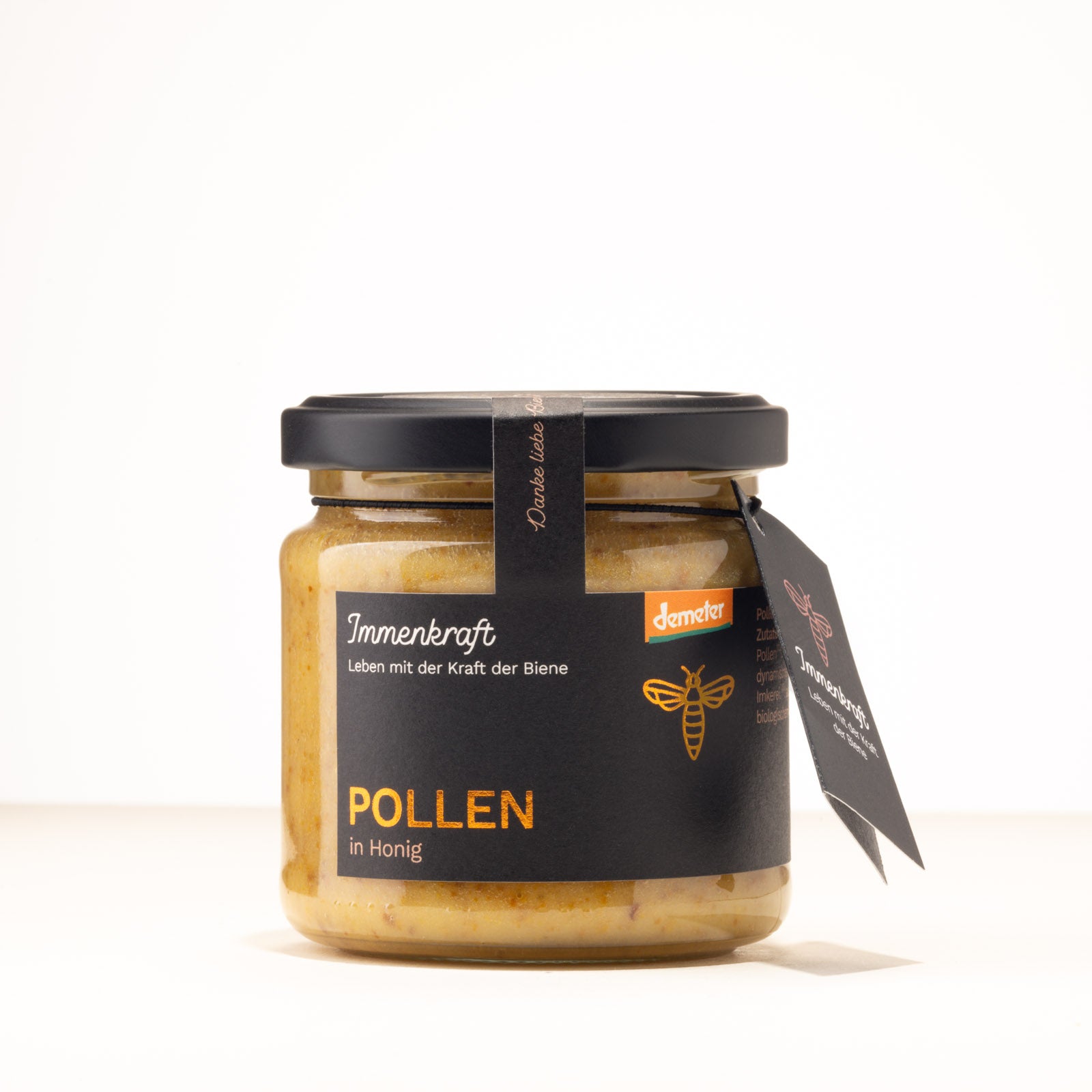 Ein 250g klares Twist-Off-Glas mit matt-schwarzem Deckel, schwarzem Anhängeetikett und Etikett mit der Aufschrift Immenkraft - Leben mit der Kraft der Biene. Produktname: Pollen in Honig. Demeter-Siegel und Bienen-Logo.